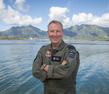 Group Captain Pete Gibson in Hawaii, stands with arms crossed and a smile in front of a body of water and hills in the background. 