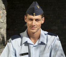 An aviator looks directly at the camera with a smile wearing his RNZAF uniform. In the background you can see a brick wall with sunlight on it and then a shadow directly behind the person.  