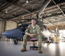 An aviator, with short dark hair wearing military uniform sits on an office chair in an aircraft hangar in front of a sleek grey helicopter.