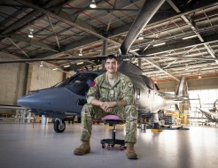 An aviator, with short dark hair wearing military uniform sits on an office chair in an aircraft hangar in front of a sleek grey helicopter.