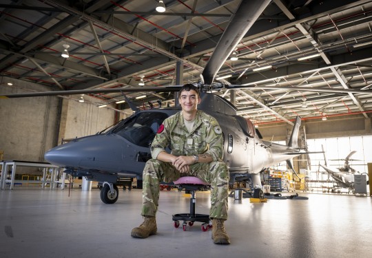 An aviator, with short dark hair wearing military uniform sits on an office chair in an aircraft hangar in front of a sleek grey helicopter.