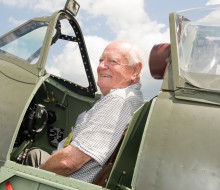 Max smiles as he sits in the open cockpit of a Spitfire aircraft on the tarmac of Base Ohakea with scattered cloud on the background.