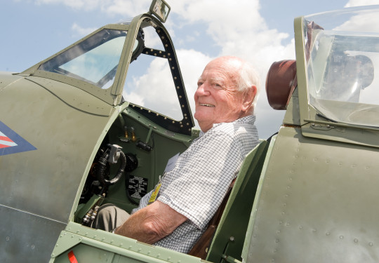 Max smiles as he sits in the open cockpit of a Spitfire aircraft on the tarmac of Base Ohakea with scattered cloud on the background.