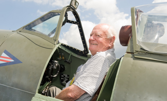 Max smiles as he sits in the open cockpit of a Spitfire aircraft on the tarmac of Base Ohakea with scattered cloud on the background.