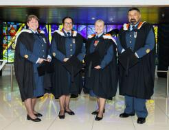 Four aviators stand in front of stained glass at the Air Force Museum of New Zealand, each in their RNZAF uniform with graduation cloaks on. 