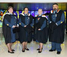 Four aviators stand in front of stained glass at the Air Force Museum of New Zealand, each in their RNZAF uniform with graduation cloaks on. 