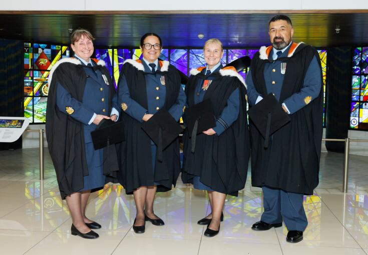Four aviators stand in front of stained glass at the Air Force Museum of New Zealand, each in their RNZAF uniform with graduation cloaks on. 