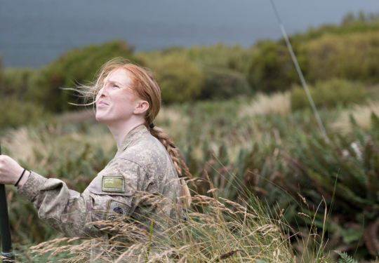 A soldier looks up while setting up a radio antenna mast in the field.