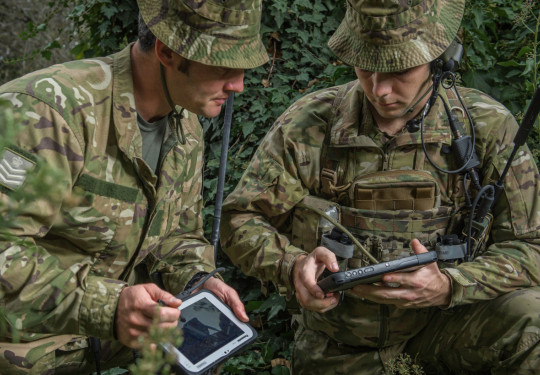 Soldiers in the field with tactical communications equipment look at tablets.