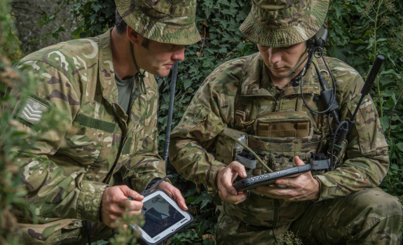 Soldiers in the field with tactical communications equipment look at tablets.