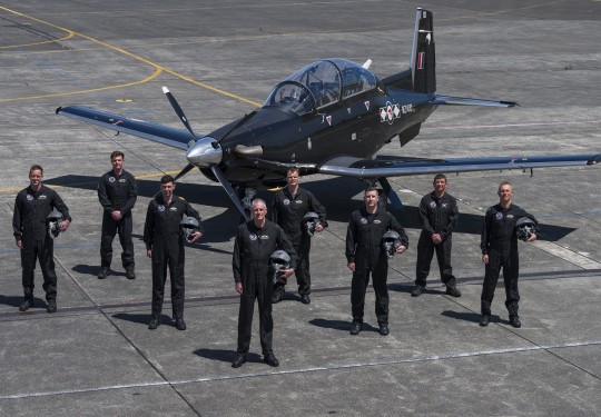 eight male pilots stand in formation in front of a small black aircraft. They each wear black flying suits and several hold flying helmets in their left arms.