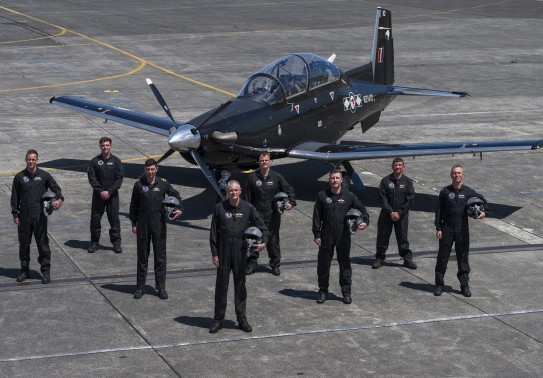 eight male pilots stand in formation in front of a small black aircraft. They each wear black flying suits and several hold flying helmets in their left arms.