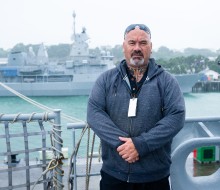 A male stands with his arms clasped in front of him, he is wearing a casual grey hoodie and jeans and a lanyard with his sunglasses on his head. He stands aboard a military naval ship and behind him is another grey ship alongside the wharf with glimpses o