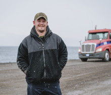 A person stands in the snow, looking and smiling at the camera. Hands in pockets and wearing a peaked cap. There is a truck in the background, along with some water.