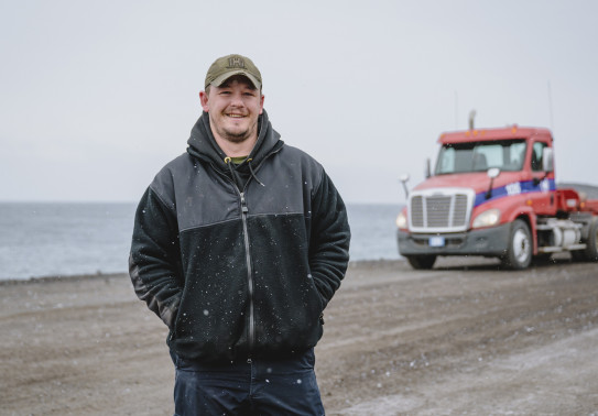 A person stands in the snow, looking and smiling at the camera. Hands in pockets and wearing a peaked cap. There is a truck in the background, along with some water.