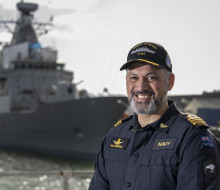 A uniformed sailor stands looking at the camera with a Navy ship in the background, out of focus. The sailor has a cap on and is smiling at the camera. 
