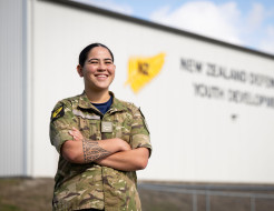 A woman with a traditional tattoo on her forearm stands with arms crossed while wearing military uniform. She smiles as she looks into the distance in front of a large grey building with words that are blurred out on the side.