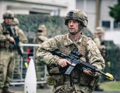 A soldier in full Army uniform and holding a weapon is in focus, while blurred in the background are more soldiers and a large bush in front of a building.