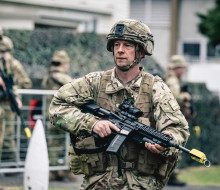 A soldier in full Army uniform and holding a weapon is in focus, while blurred in the background are more soldiers and a large bush in front of a building.