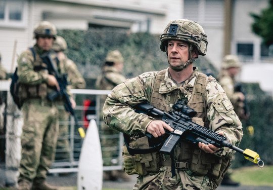 A soldier in full Army uniform and holding a weapon is in focus, while blurred in the background are more soldiers and a large bush in front of a building.