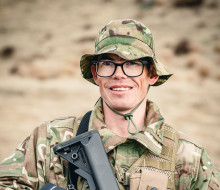A soldier stands looking at the camera holding a weapon, in uniform