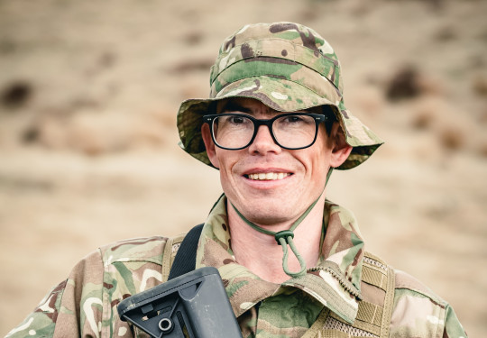 A soldier stands looking at the camera holding a weapon, in uniform