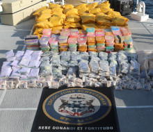 A large pile of seized drugs photographed on the deck of USCGC John Scheuerman. The packaged drugs range from small clear plastic bags (front) to larger coloured tupperware, with large yellow bags in the background.
