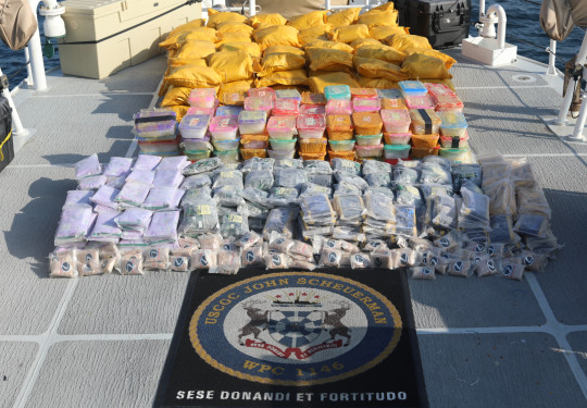 A large pile of seized drugs photographed on the deck of USCGC John Scheuerman. The packaged drugs range from small clear plastic bags (front) to larger coloured tupperware, with large yellow bags in the background.