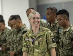 A blonde lady with glasses in an Army uniform smiles in front of a group of Malaysian Armed Forces soldiers.
