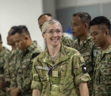 A blonde lady with glasses in an Army uniform smiles in front of a group of Malaysian Armed Forces soldiers.