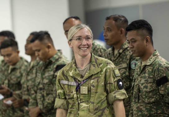 A blonde lady with glasses in an Army uniform smiles in front of a group of Malaysian Armed Forces soldiers.