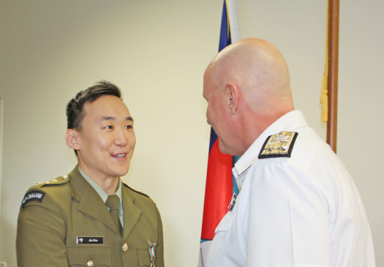 Captain Jin Cha in military dress uniform, receives the New Zealand Defence Service Medal from Commander Joint Forces New Zealand, Rear Admiral Jim Gilmour. The pair shake hands.