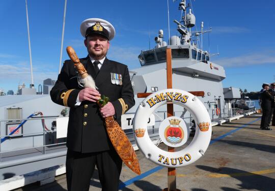 A Royal New Zealand Navy sailor holds a symbol of command in front of a grey navy ship under blue skies.