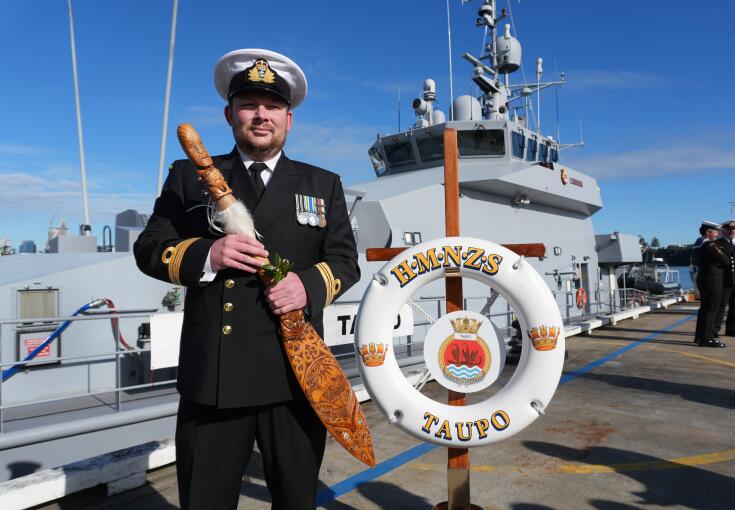 A Royal New Zealand Navy sailor holds a symbol of command in front of a grey navy ship under blue skies.