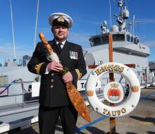 A Royal New Zealand Navy sailor holds a symbol of command in front of a grey navy ship under blue skies.