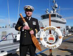 A Royal New Zealand Navy sailor holds a symbol of command in front of a grey navy ship under blue skies.
