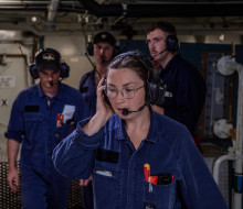 A woman wearing Navy blue overalls, glasses and a headset at work in the electrical part of a ship.