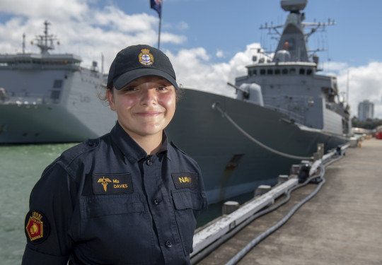 A sailor stands smiling on a sunny day, with some clouds. There are two ships in the background. 