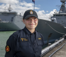 A sailor stands smiling on a sunny day, with some clouds. There are two ships in the background. 