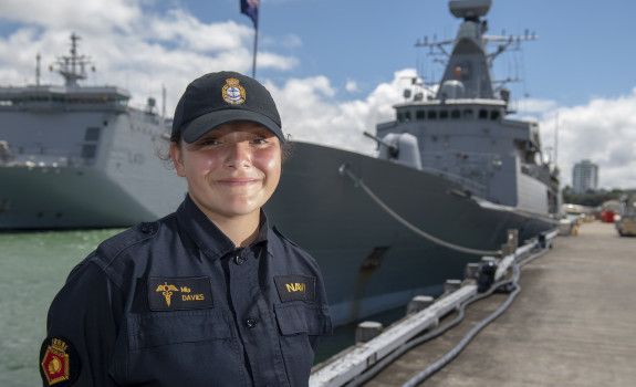 A sailor stands smiling on a sunny day, with some clouds. There are two ships in the background.