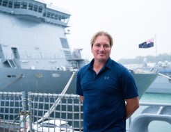 A blonde haired, middle aged man stands in front of two large grey naval ships.  He wears a navy blue under armour polo shirt and stands casually with his arms behind his back. Behind him the New Zealand flag flies prominently on the bow of the closer shi