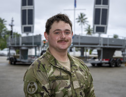 A man with a mostache in military uniform looks at the camera as he stands in front of the two grey USVs on trailers.