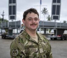 A man with a mostache in military uniform looks at the camera as he stands in front of the two grey USVs on trailers.