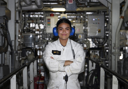 Sub Lieutenant Emily Aull stands in an engine room with arms crossed, wearing blue ear protectors and white overalls with a black and gold name badge.