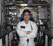 Sub Lieutenant Emily Aull stands in an engine room with arms crossed, wearing blue ear protectors and white overalls with a black and gold name badge.