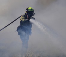 A firefighter emerges from the smoke with their hose on as it continues to spray water. 