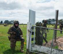 Officer cadets are spaced out along a wire fence in rural Stratford. They are wearing camouflage, vests, helmets and weapons. 