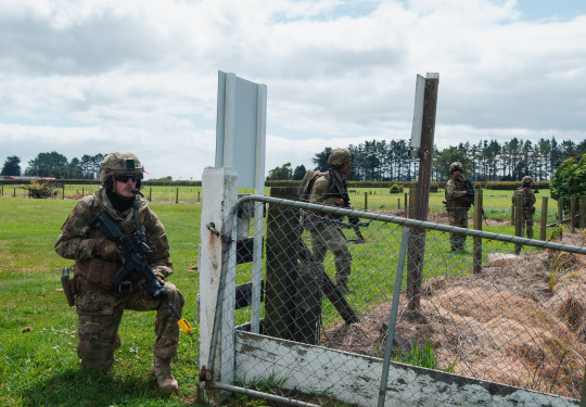 Officer cadets are spaced out along a wire fence in rural Stratford. They are wearing camouflage, vests, helmets and weapons. 