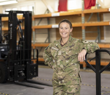 Flying Officer Challies-Kolk standing in a warehouse leaning on something. There is a forklift in the background.. 