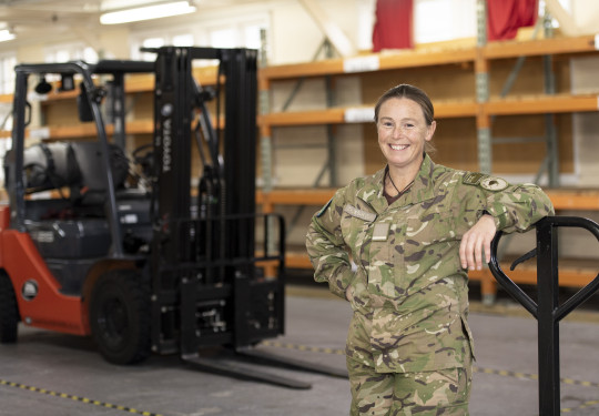 Flying Officer Challies-Kolk standing in a warehouse leaning on something. There is a forklift in the background.. 